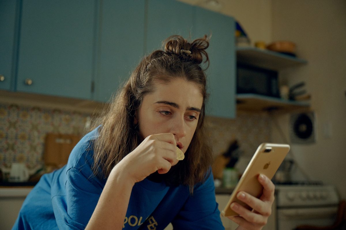 Sam est abaissée vers une table en regardant attentivement son portable tout en mangeant un biscuit. Elle est visiblement perturbée par ce qu'elle lit.