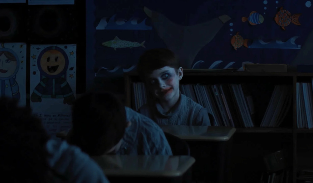 Avec un maquillage de clown un peu cliché mais néanmoins efficace, un enfant, le seul à être levé au milieu de ses camarades, regarde très fixement la caméra, un grand sourire aux lèvres.