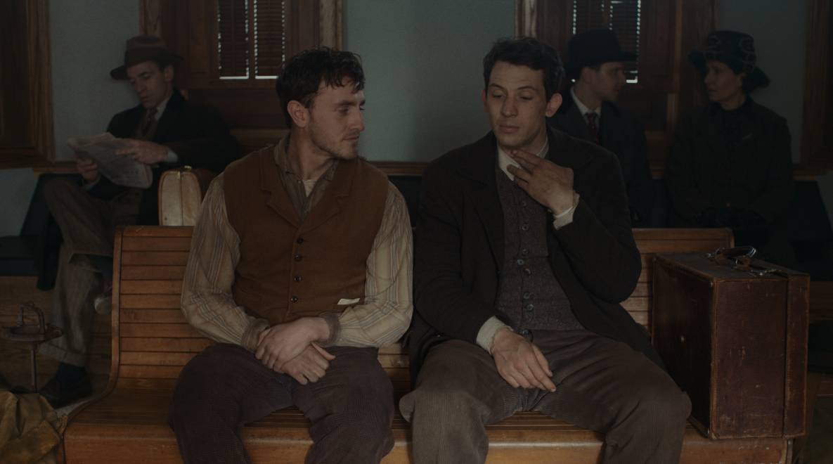 Image du film Le son des souvenirs. David et Lionel sont assis sur un banc d'une gare de province. David explique à Lionel une technique vocal en se touchant les cordes vocales tandis que ce dernier le regard tendrement.