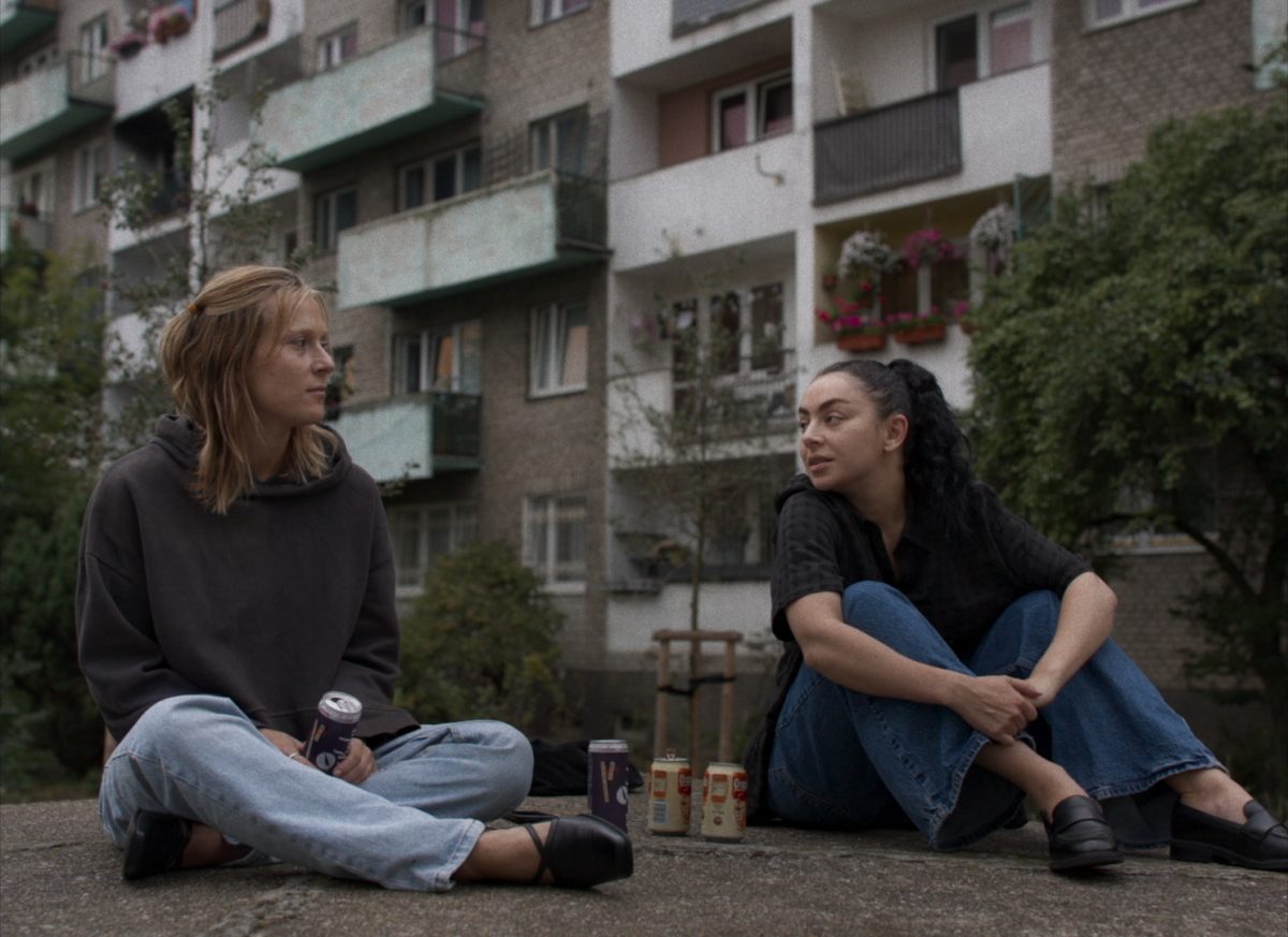 Image issue du film Eruption. Nel et Bethany sont assises l'une à côté de l'autre sur ce qui s'apparente à un rocher et font dos à un immeuble. Elles se regardent attentivement et semblent en pleine conversation intense. 