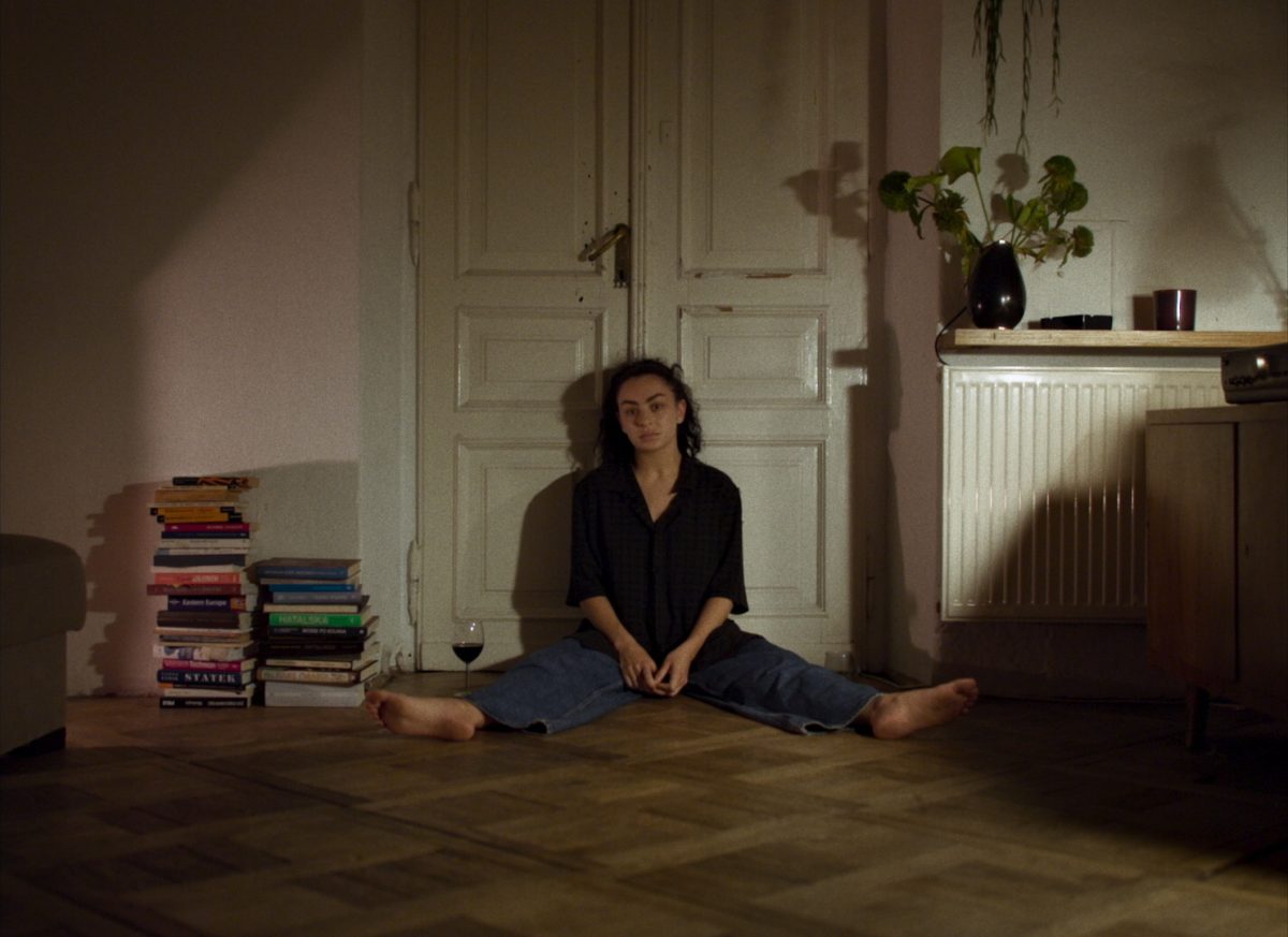 Image issue du film Eruption. Bethany est assise par terre, chez Nel, dos contre une double porte et regarde la caméra directement visiblement perdu dans ses réflexions internes. 
