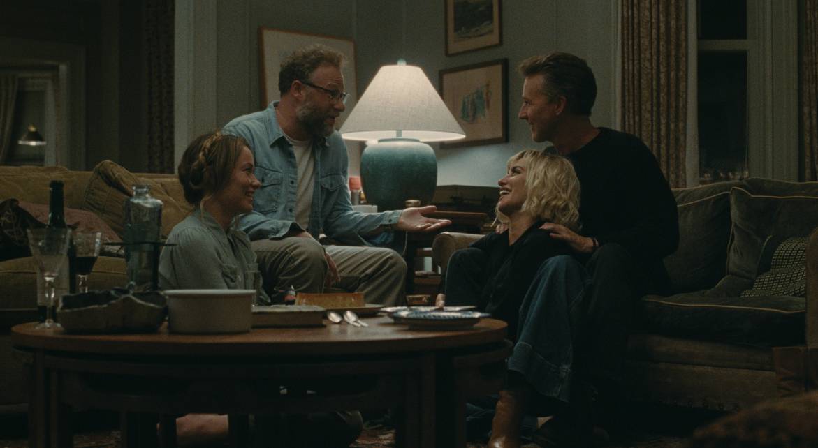 Photo de L'Invitation. Les deux couples (Angela et Joe - Piña et Hawk) se font face dans la salon des protagonistes. Les hommes sont assis sur le canapé et les femmes par terre, entre les jambes de leur conjoint. Ils semblent rigoler et vivre un moment léger.